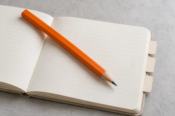 An orange pencil lies across a neatly organised cream notebook with blank index tabs.