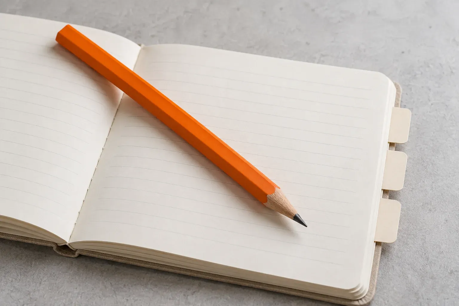 An orange pencil lies across a neatly organised cream notebook with blank index tabs.